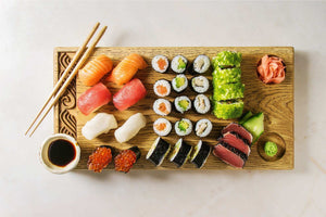 Overhead view of assorted sushi, sashimi, and nigiri arranged on a wooden serving board with chopsticks, soy sauce, pickled ginger, and wasabi