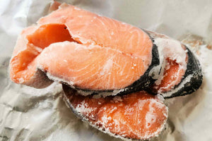 Close-up overhead of frozen salmon steaks showing bright orange flesh with ice crystals and skin on, resting on crumpled foil