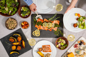 Overhead table with lemon-garnished roasted salmon on a slate platter, bowls of salad, roasted vegetables, quinoa, cheese and glasses of white wine.