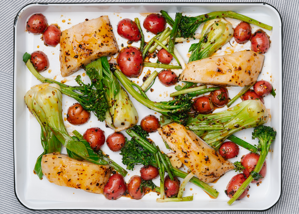 Baked fish fillets glazed with chili oil alongside baby red potatoes and broccolini in a white baking tray on a striped cloth