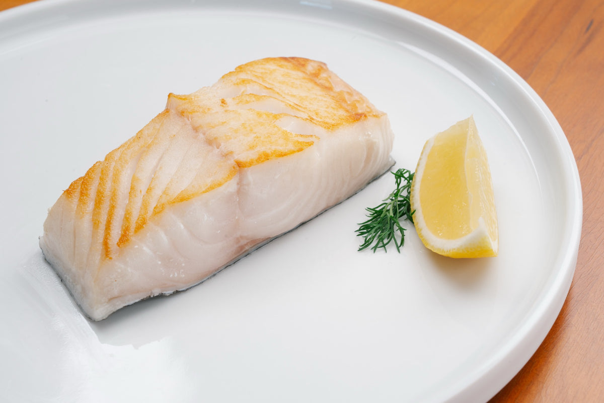 A seared black cod fillet on a white plate with a lemon wedge and dill