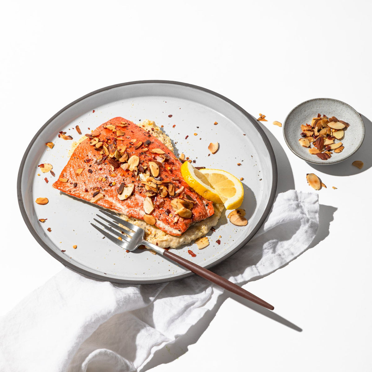 A seared wild salmon fillet topped with toasted sliced almonds and a lemon wedge on a white plate, with a fork and linen napkin