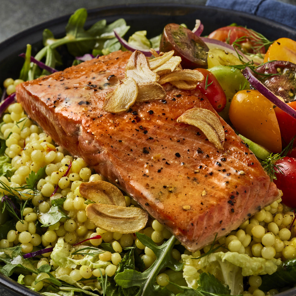 A seared wild salmon fillet with toasted garlic on pearl couscous with greens, tomatoes and red onion in a dark bowl