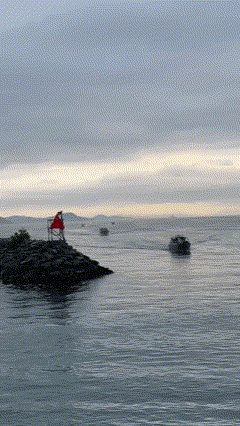 Calm grey harbour water with a red beacon on a rocky point and boats in the distance