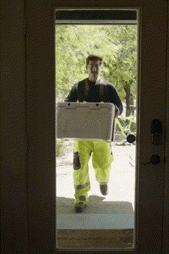 A man carrying a white cooler up to a glass front door, seen from inside the house